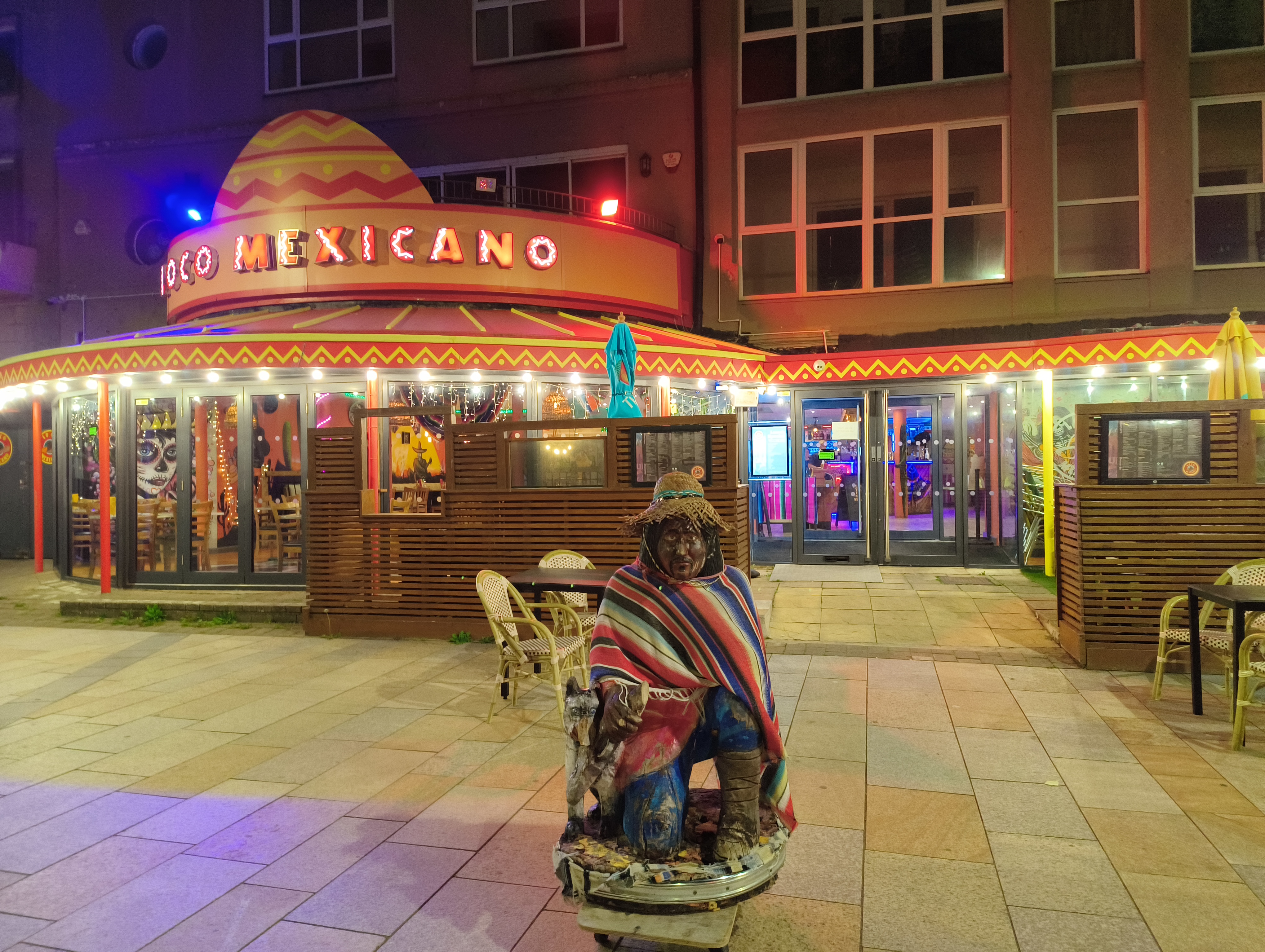 The brightly lit exterior of the "Loco Mexicano" restaurant, featuring a large, yellow-domed sign and a colorful, kneeling statue of a person in a sombrero and serape in the foreground.
