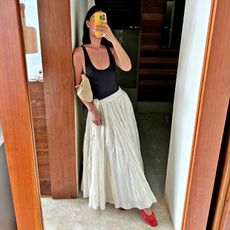 fashion influencer Mimi Nguyen poses for a mirror selfie while on vacation in Tulum Mexico with a bright yellow iphone case wearing a low-cut black tank top, tan suede bag as a clutch, full white maxi skirt, and red mesh flats