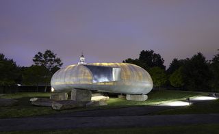 Alternative exterior view of the light grey, curved and illuminated 2014 Serpentine Pavilion by Smiljan Radic at night. The structure sits on large stones and is surrounded by greenery. The pathway outside the structure is partially lit up by lighting in the ground