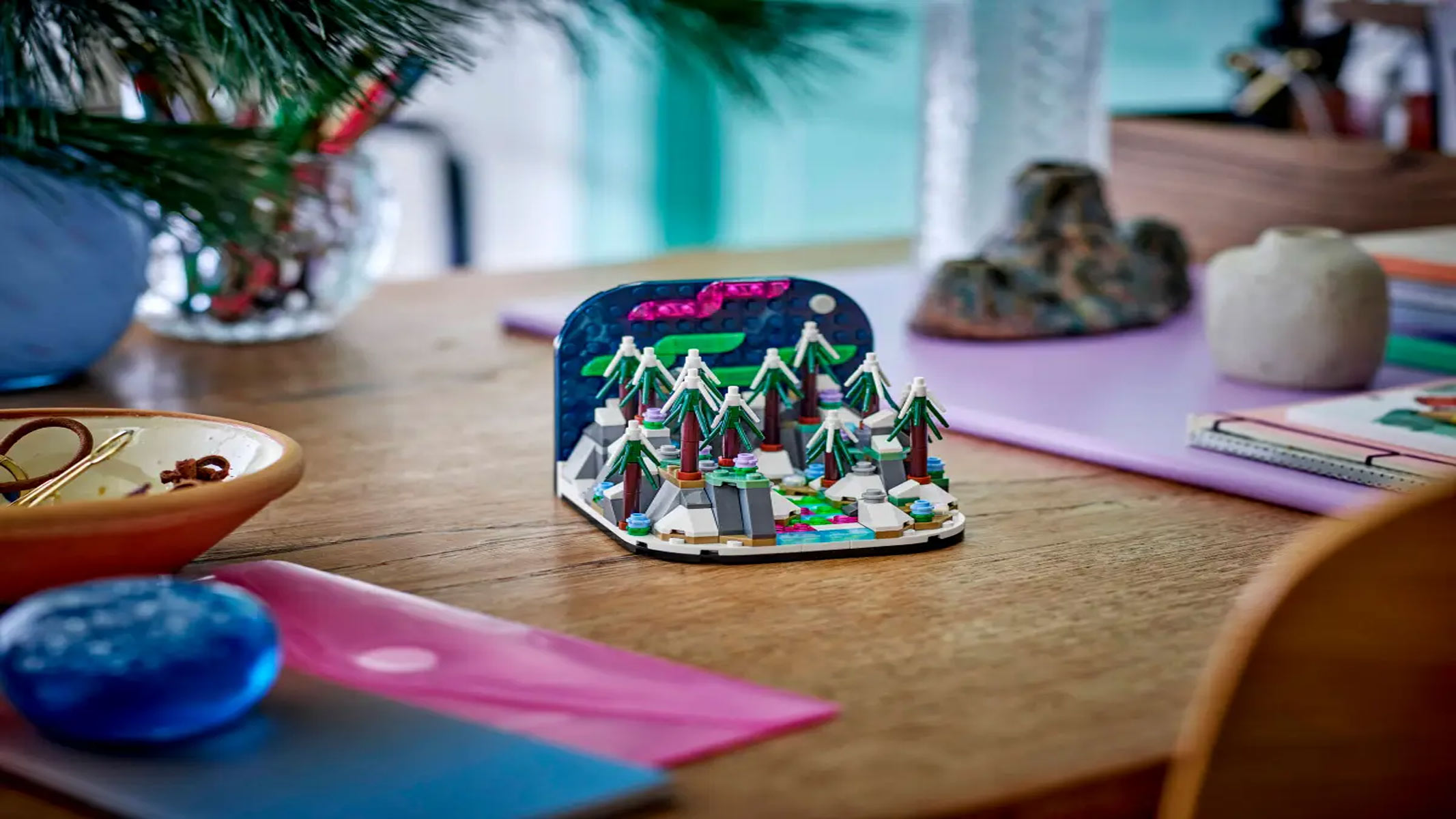 A brick-built depiction of a wintery landscape with snowy trees and running water shows the Northern Lights, on a wooden table with vibrant décor surrounding it.