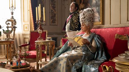 Production still of Netflix's Bridgerton series where Queen Charlotte is sitting on a red sofa in a regal blue dress. There are lots of antique side tables around here with candelabras on them.