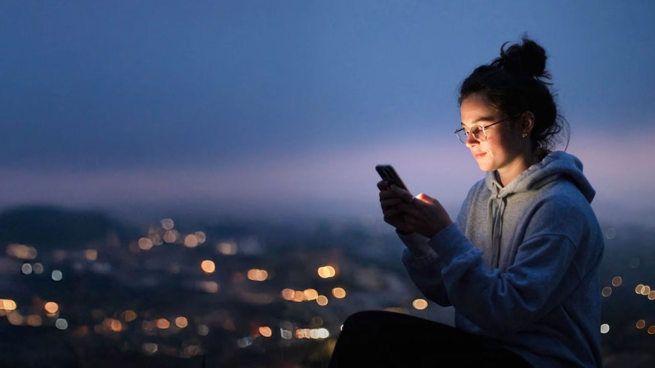 A woman is looking at her mobile phone while wearing a hoodie in front of a city skyline at dusk.