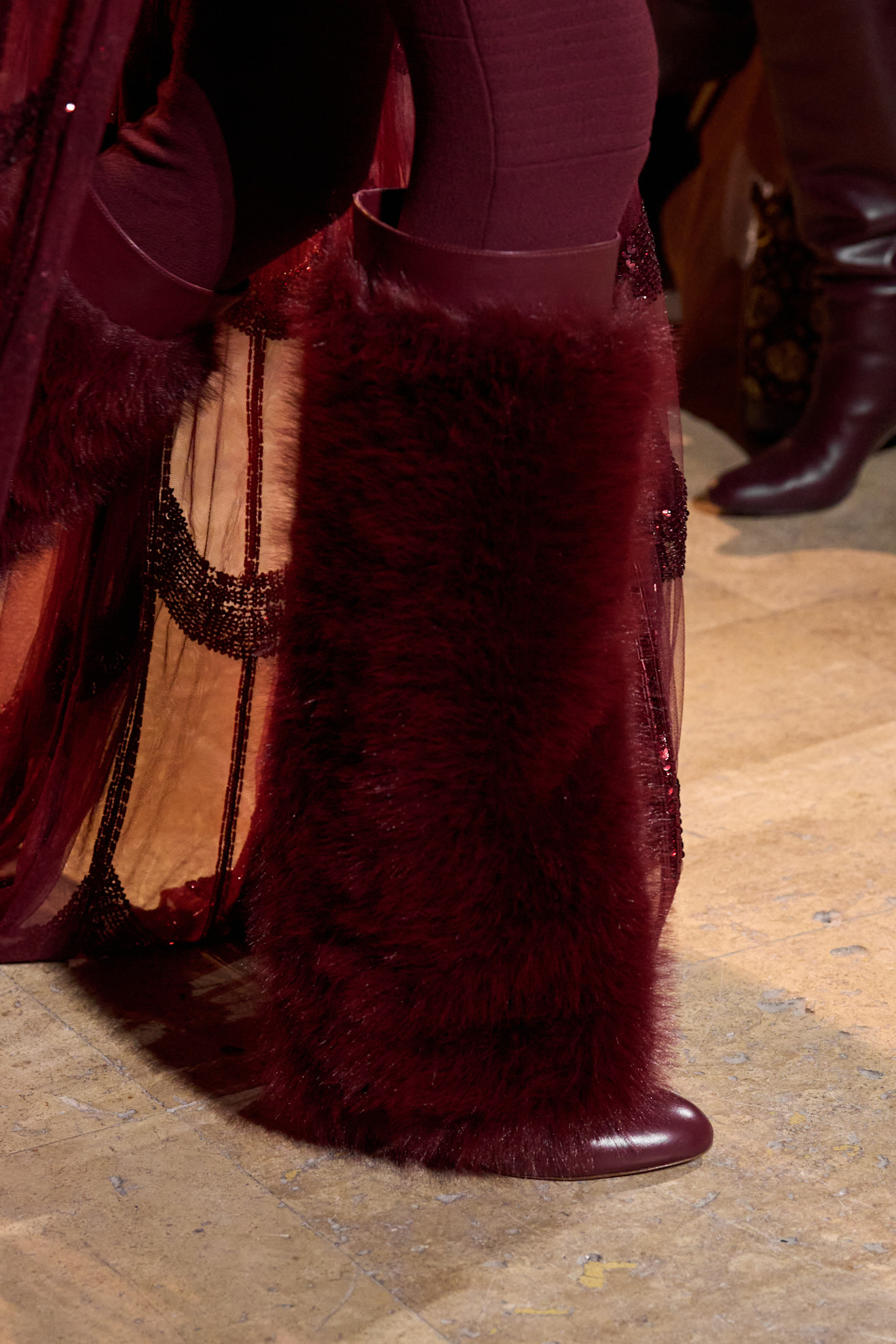 Image from the autumn/winter 2025 Elie Saab runway. The model wears a knee-high faux fur boot in burgundy.