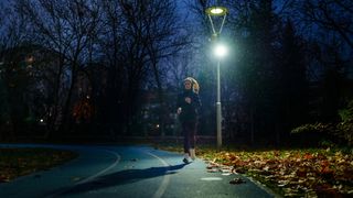 A woman jogging past a streetlamp in a park on a dark winter morning