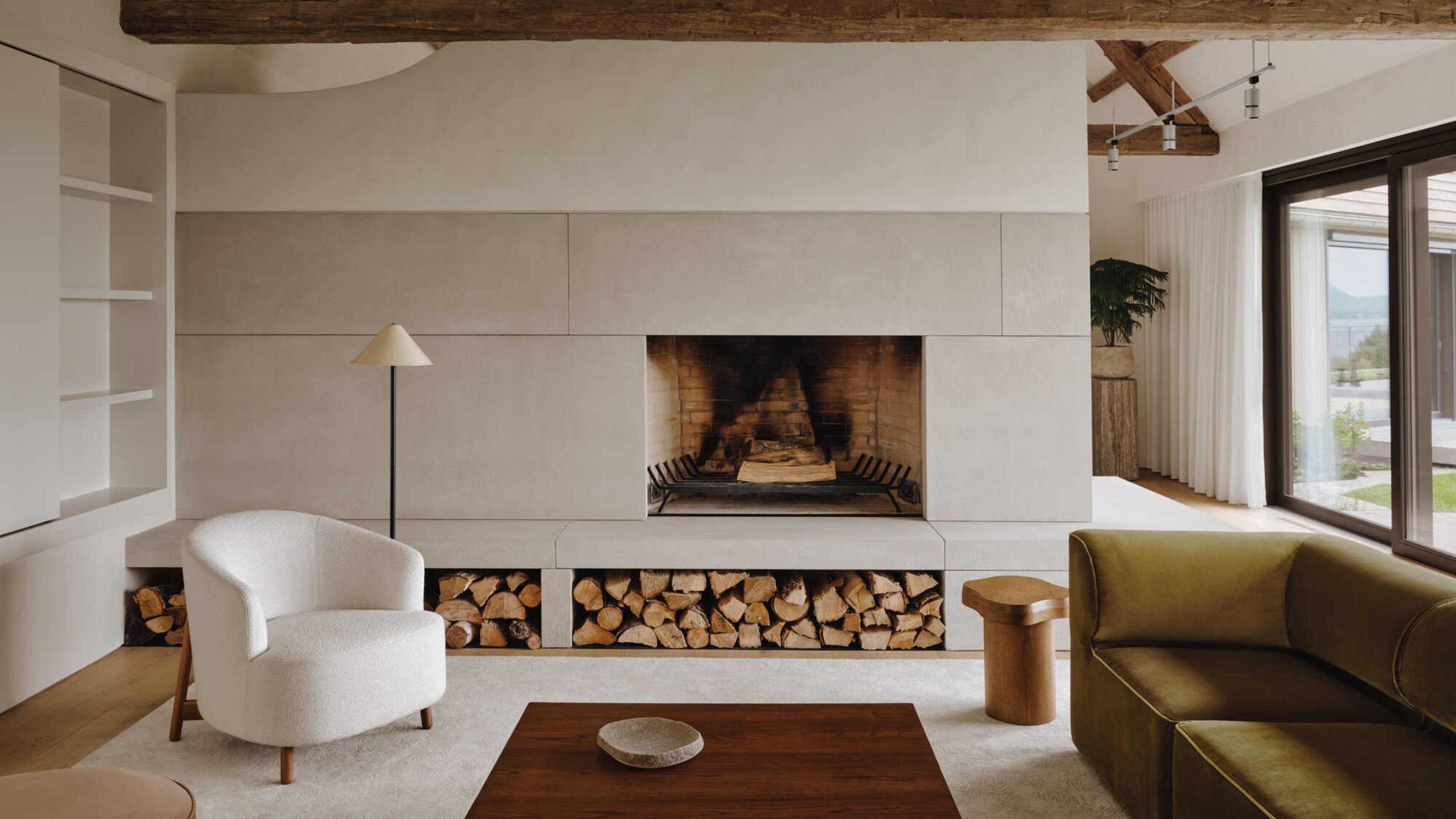 modern living room with built-in fireplace with logs stacked, a white armchair, green sofa, and exposed timber ceiling beams