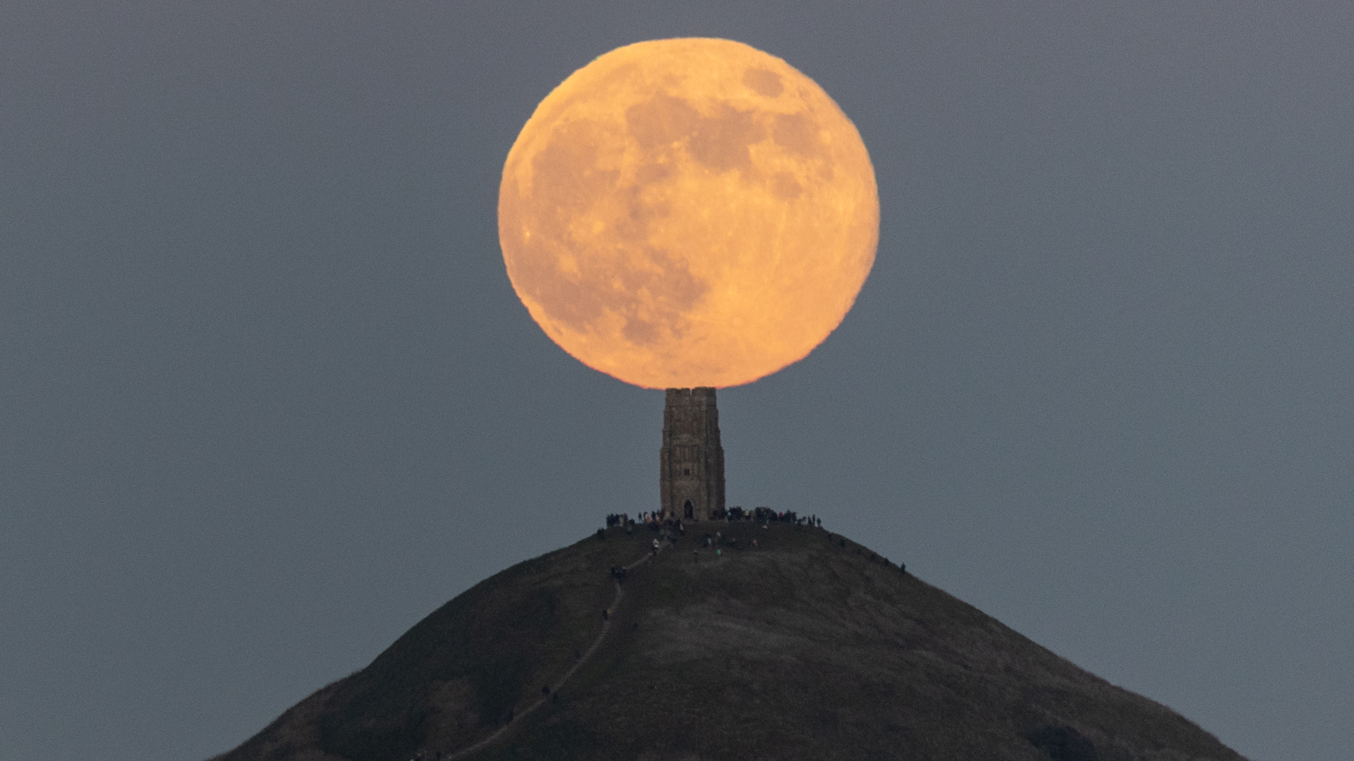 A full moon is pictured rising in a darkening blue evening sky above a lonely tower on a hill, as people gather on the hilltop to watch the event.