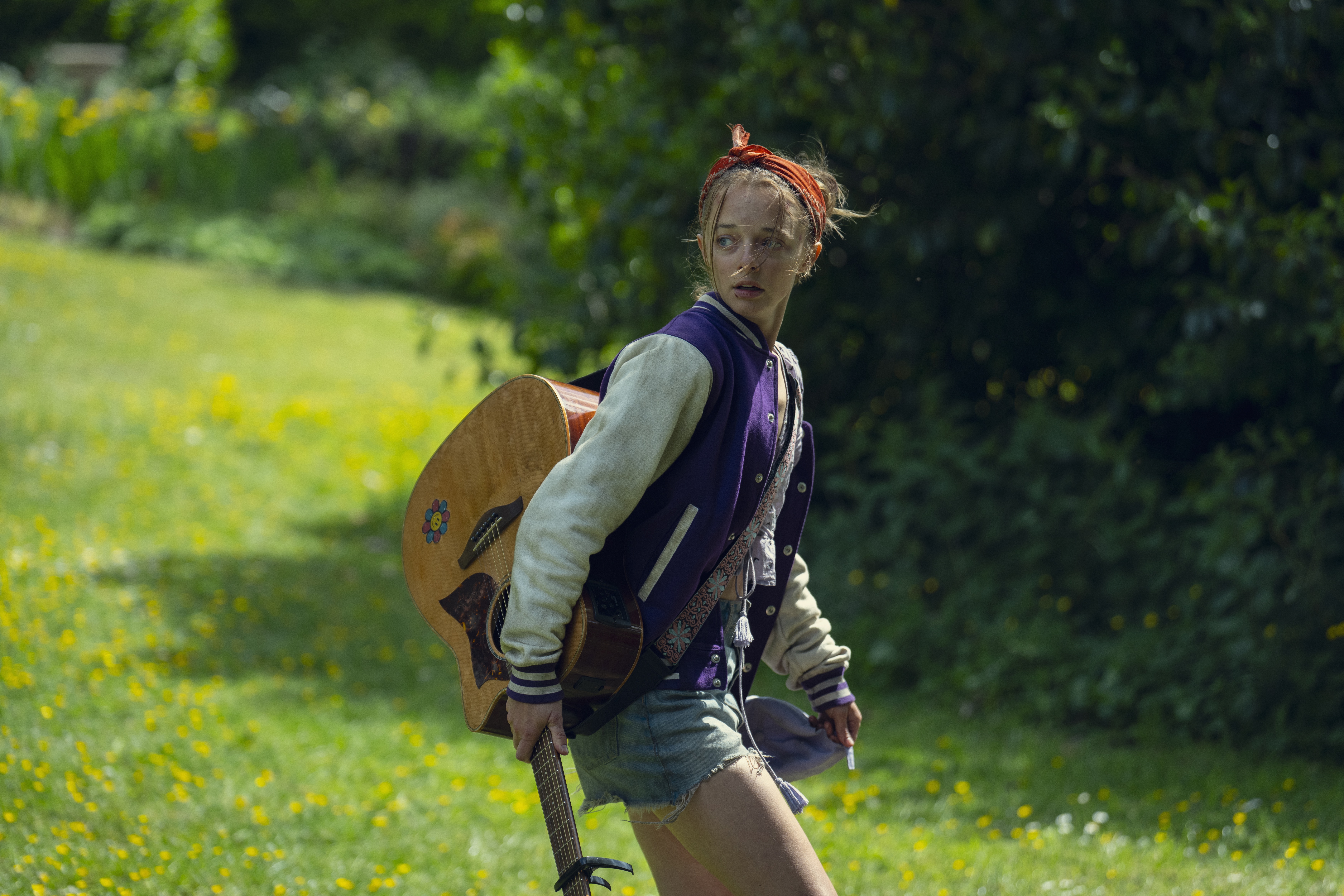 Elle de Lange as paige green looking disheveled walking through a field in shorts a letterman jacket and carrying an acoustic guitar in a still from netflix's Run Away