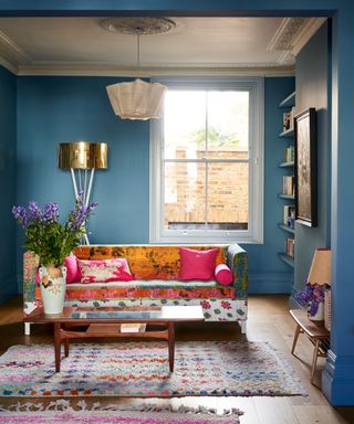 Mid blue living room with bright window, alcove shelving, brightly colored fabric soda, multi colored rugs, mid century coffee table and vase of purple flowers