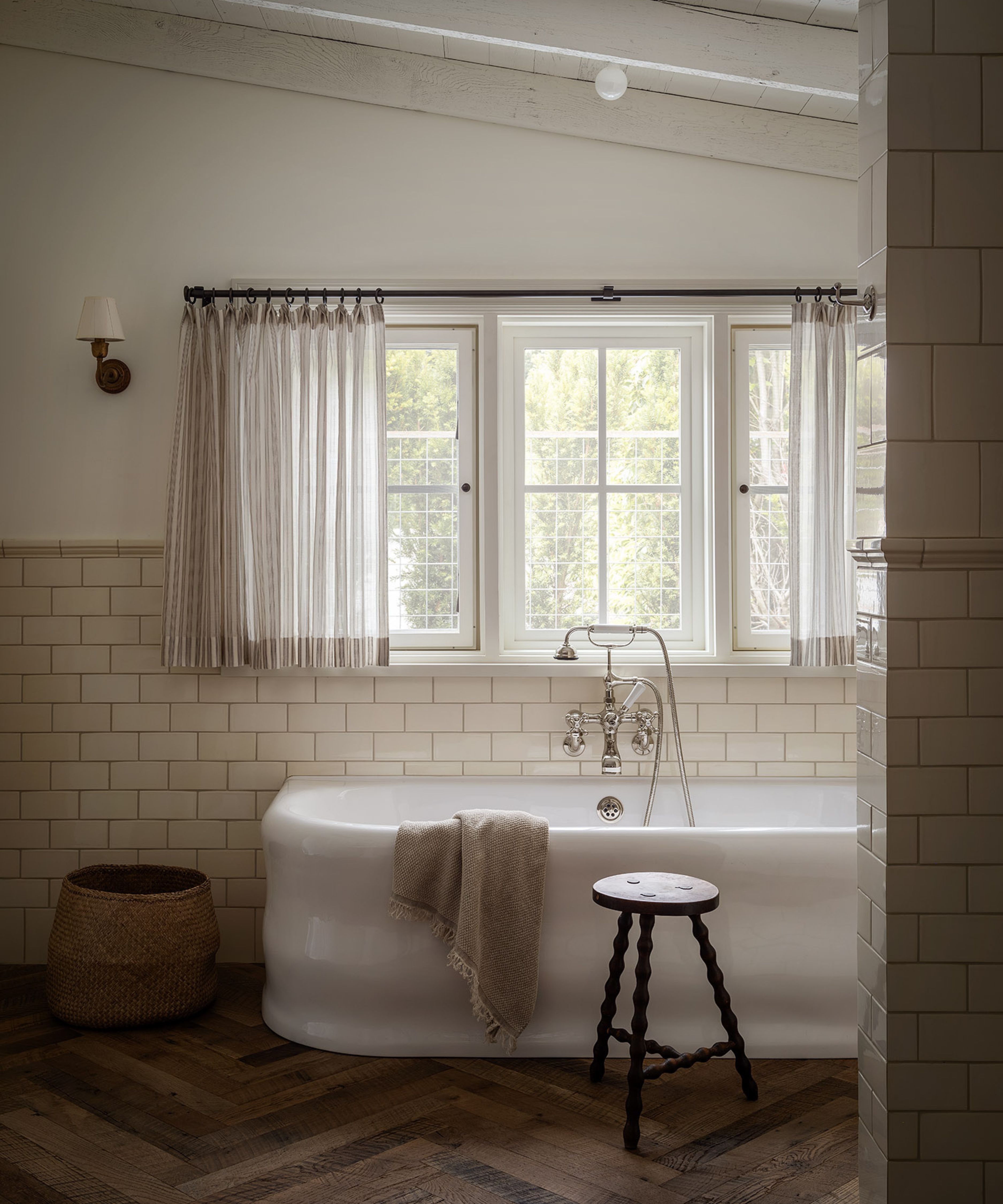 a warm white bathroom with white painted ceiling beams and a herringbone wooden floor, a freestanding bathtub sits beneath a large window with drapes and a small wall sconce