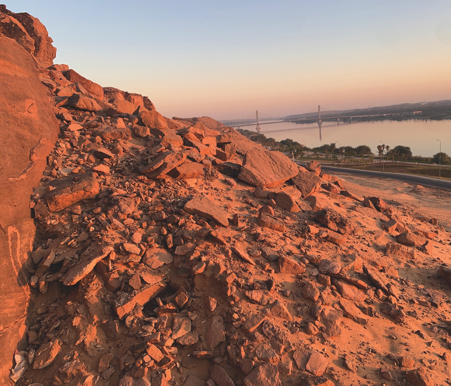 a view of a rocky hill with a road, a body of water and bridge visible in the distance
