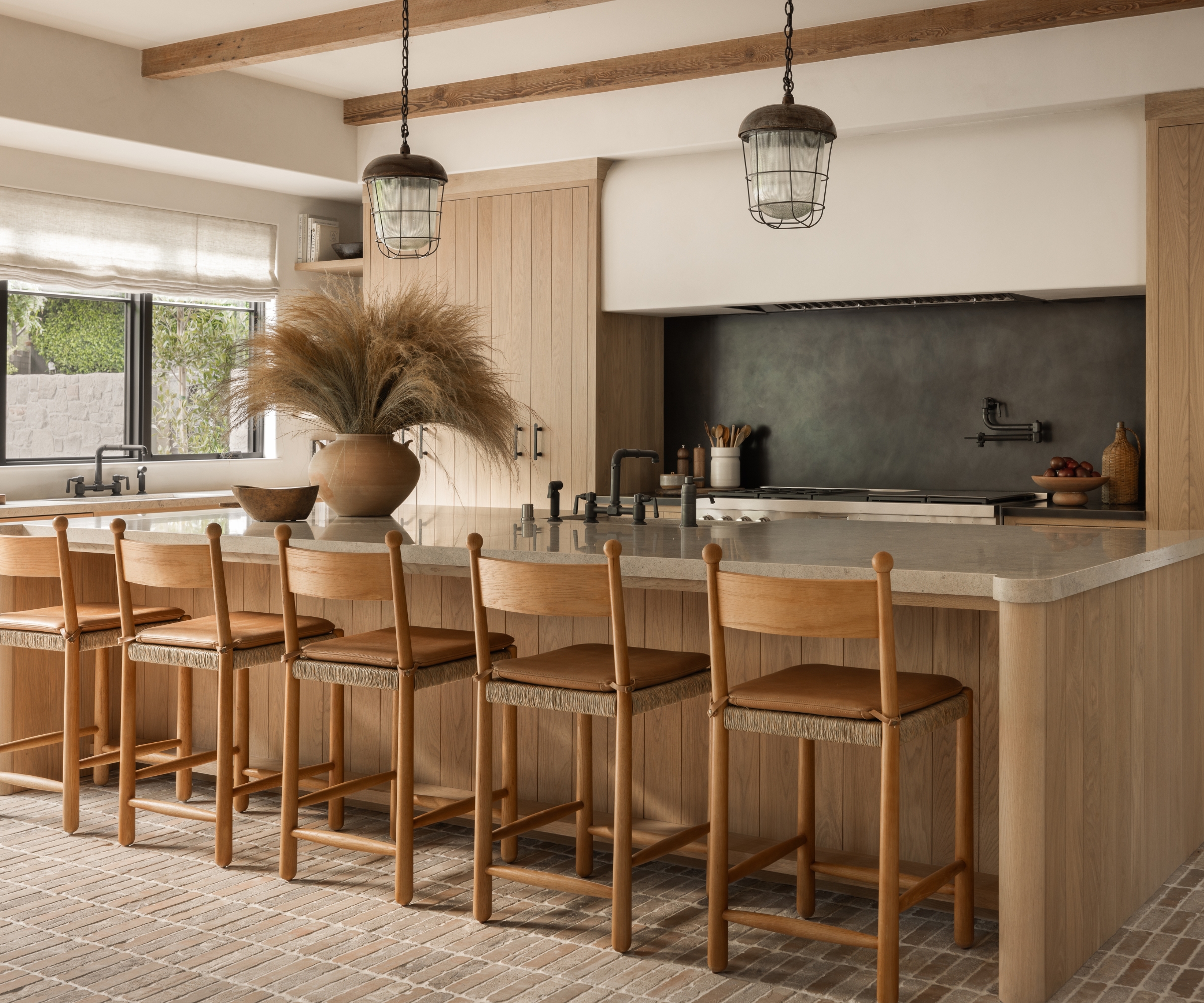 A large wooden kitchen with shiplap-inspired cabinets, a black backsplash, brick flooring, and industrial hardware