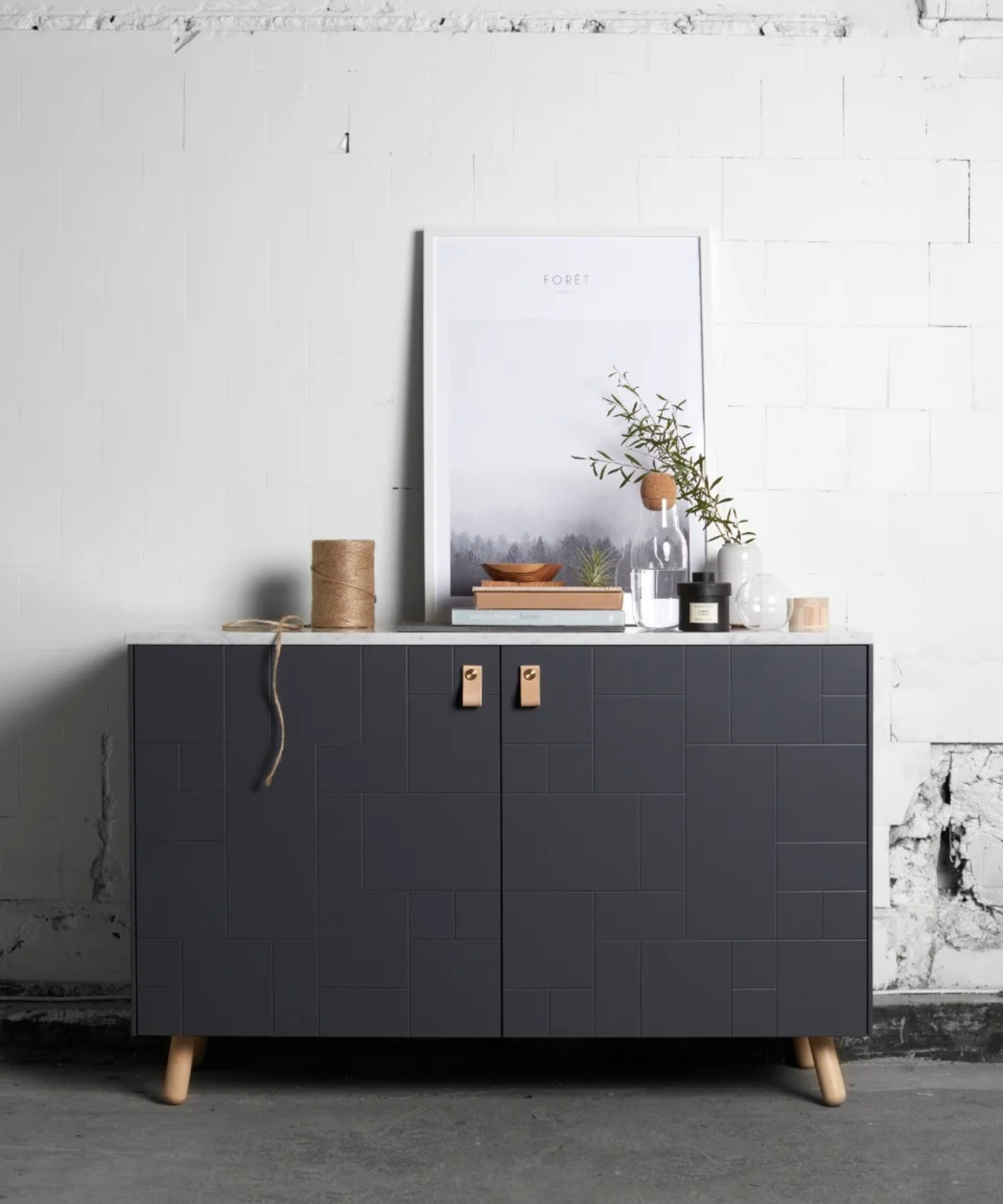 sideboard with two grey doors with leather pull handles and wooden angled legs against white distressed finish wall