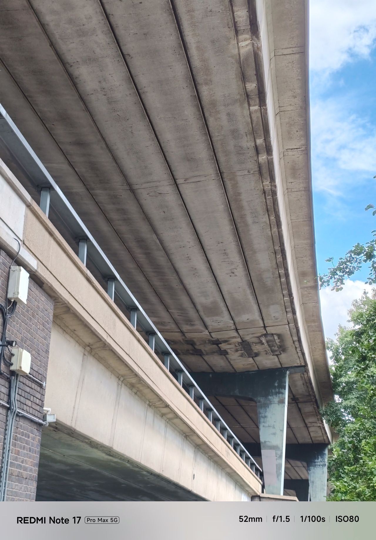Upward view beneath a concrete elevated roadway with blue sky and trees visible alongside it.