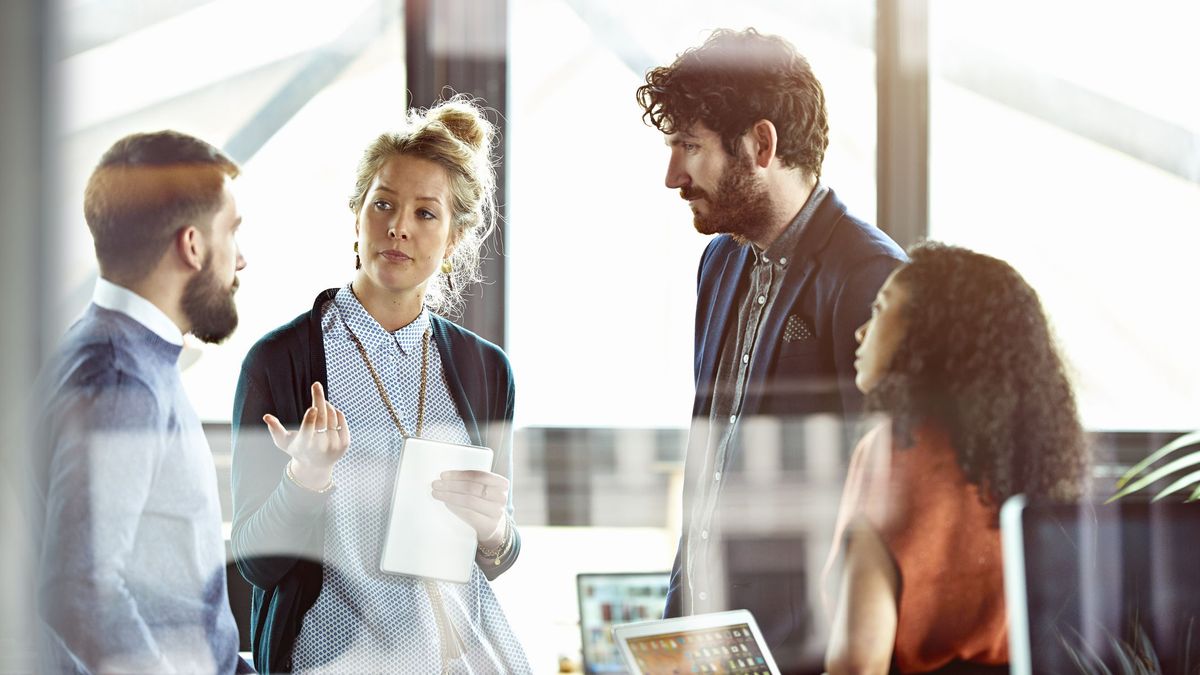 Two men and a woman in business attire listen to a third businesswoman talk, while the man and woman on the right hold laptops. A reflection partially obscures the man and woman on the left of the frame.