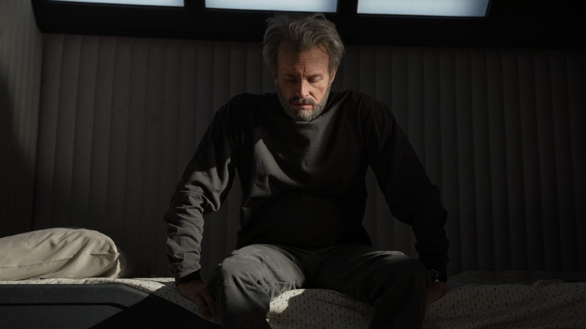 In a very plain, gray room there is a worried older man sitting on a white bed looking down. He has dishevelled short, gray hair, as well as a gray beard and moustache. He is wearing a dark gray long-sleeved shirt and trousers.