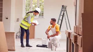 woman wearing hi vis vest and white hard hat with clipboard in hand talking to male decorator inside a room being decorated