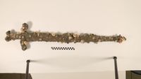 A barnacle and sand encrusted sword hangs on a white wall. 