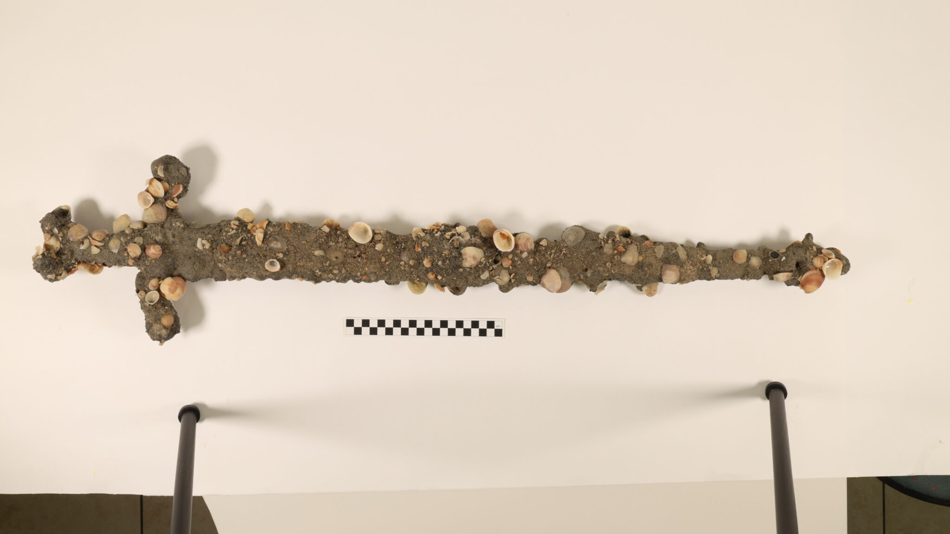 A barnacle and sand encrusted sword hangs on a white wall. 