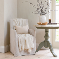 White chair and chair skirt in the corner of a neutral room, next to a marble side table with vase.