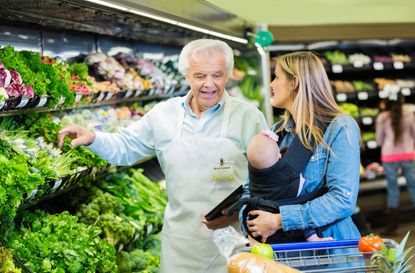 Senior adult Caucasian man with gray hair is supermarket employee or produce manager. Man is assisting customer who is shopping for healthy food and fresh vegetables. Customer is mid adult Ca