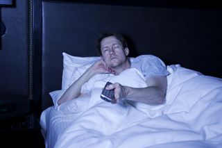 Photo of a man lying in bed watching late night television with remote control in hand with an utterly exhausted look on his face