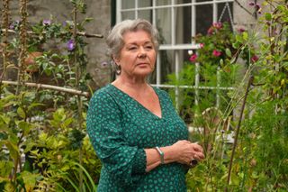 Anne Lloyd (Barbara Flynn) stands in the garden outside her cottage, wearing a green dress and looking pensive