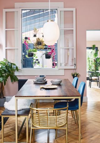 Pink dining room with internal window into kitchen, brass and walnut table and bench, and blue and brass chairs
