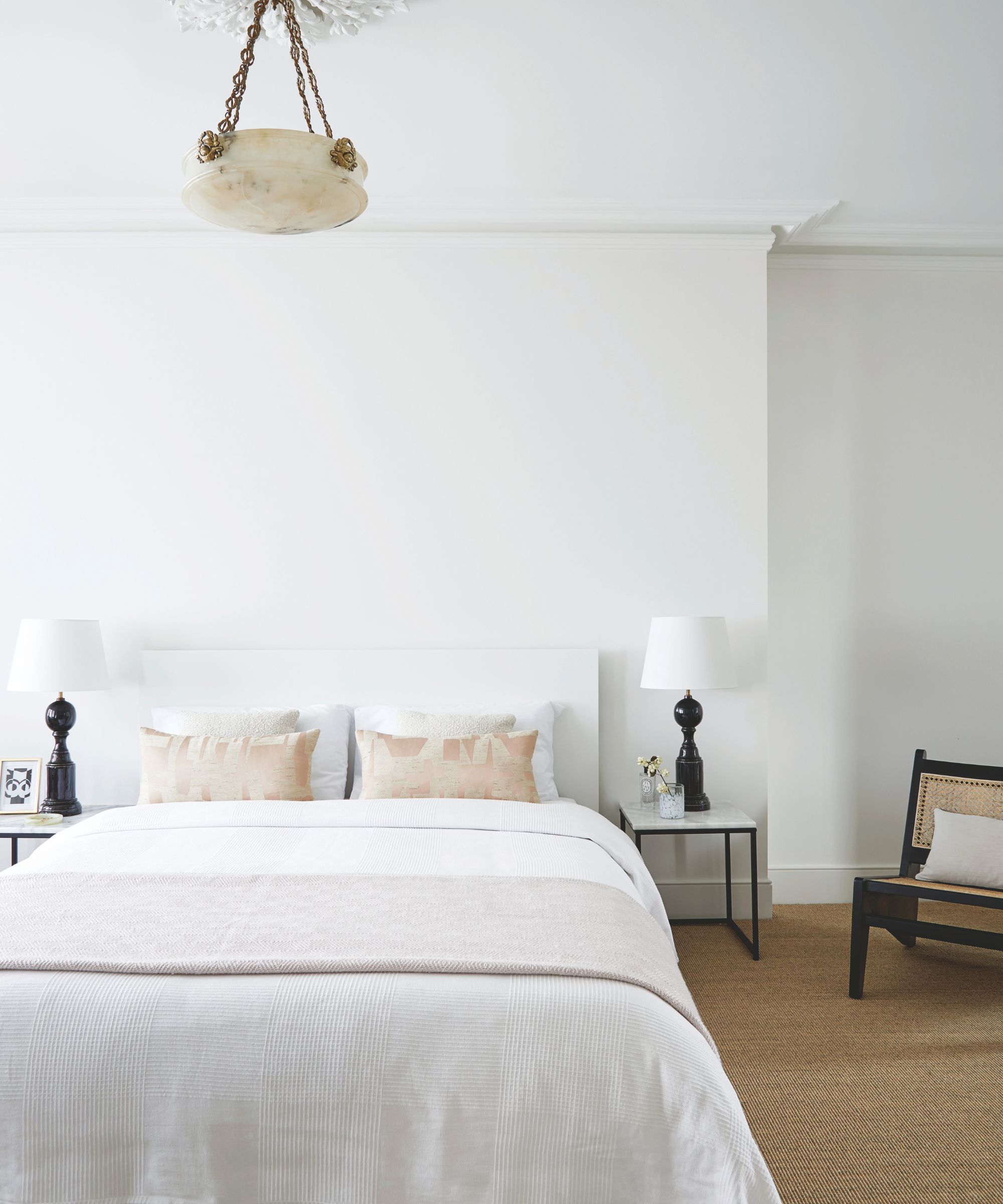Bedroom with white walls, a white bed with white bedding, two nightstands with black and white table lamps, and a jute carpet with a midcentury rattan chair in the corner.