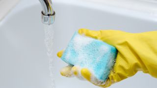 hand wearing rubber gloves holding a spone with water running from a tap in the sink