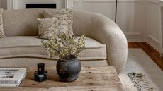 A curved boufle sofa in a cream living room, a rustic wooden coffee table in front and a rattan lamp behind