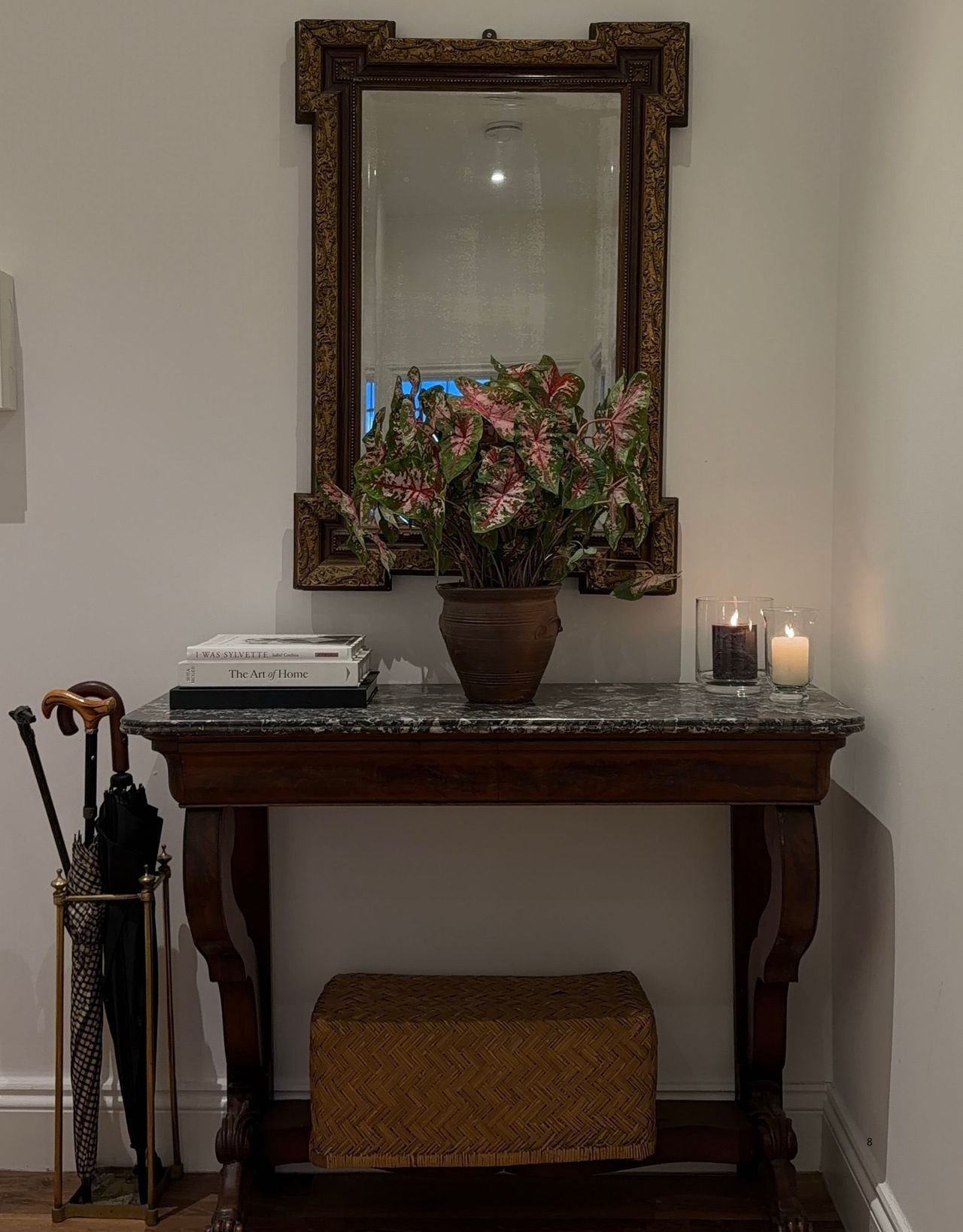 Image of a white entryway with a dark wooden table with marble on top. There is an antique mirror over it, a vintage umbrella stand to the left, and candles, a vase of flowers, and magazines on it.