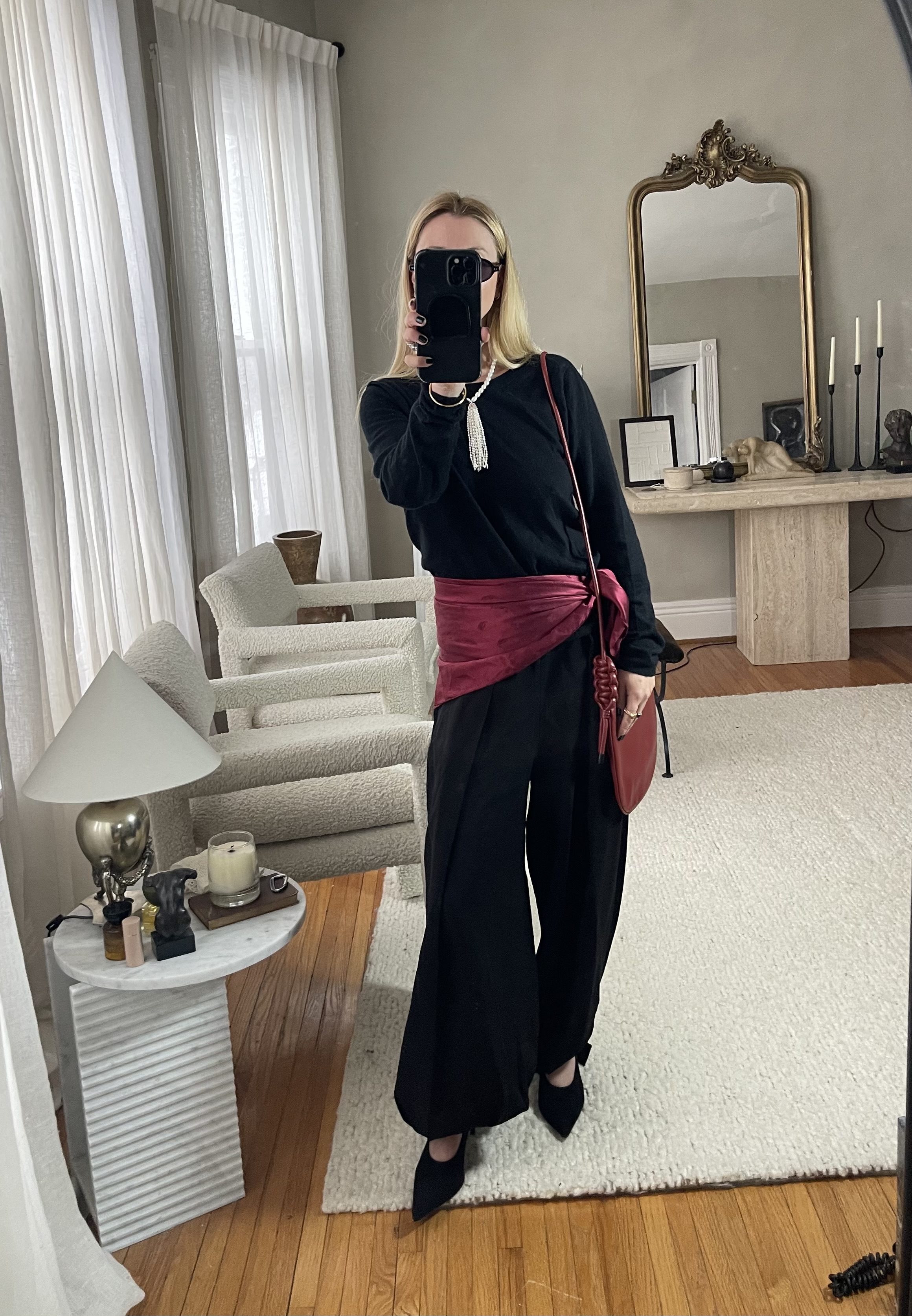 Kerry Pieri wearing a black top, black pants, a red scarf as a blet, red handbag, black heels, and a white tassel necklace