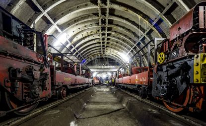 Royal mail underground train network, arched girder roof with lighting, two rail tracks with red royal mail postal trains stationed, middle concrete sectioned pathway in-between the rail tracks