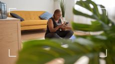 Woman scrolling on phone sitting on a yoga mat at home, surrounded by plants after finishing a Pilates workout, learning about what is Lagree
