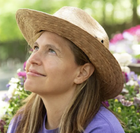 Woman with long hair wearing a strew hat and a blue top in front of flowers