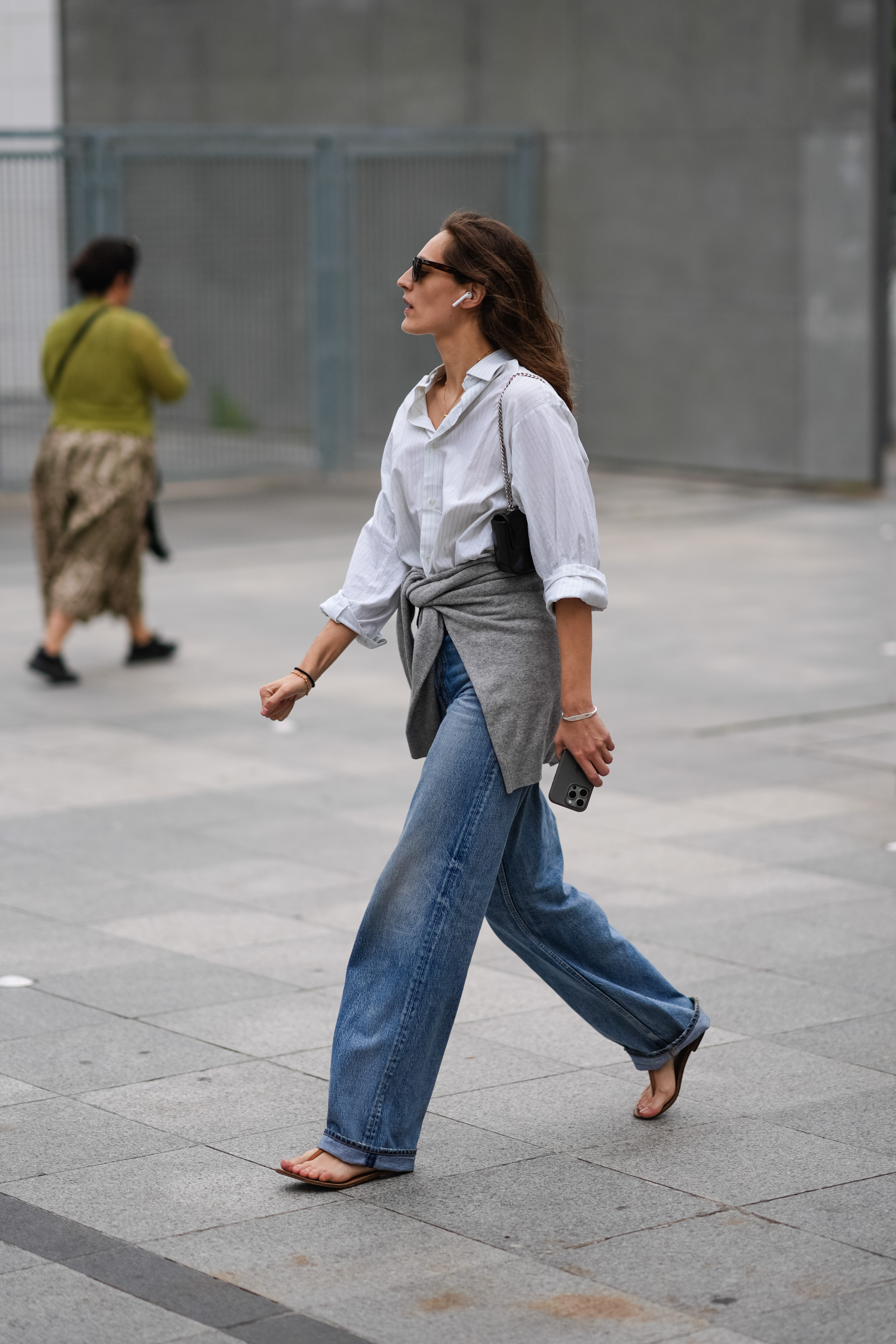 A guest wears black sunglasses, white button up long sleeve shirt, washed blue straight denim jean pants, gray sweater wrapped around waist, silver bracelet, brown thong sandal leather shoes, outside 032C, during the Paris
