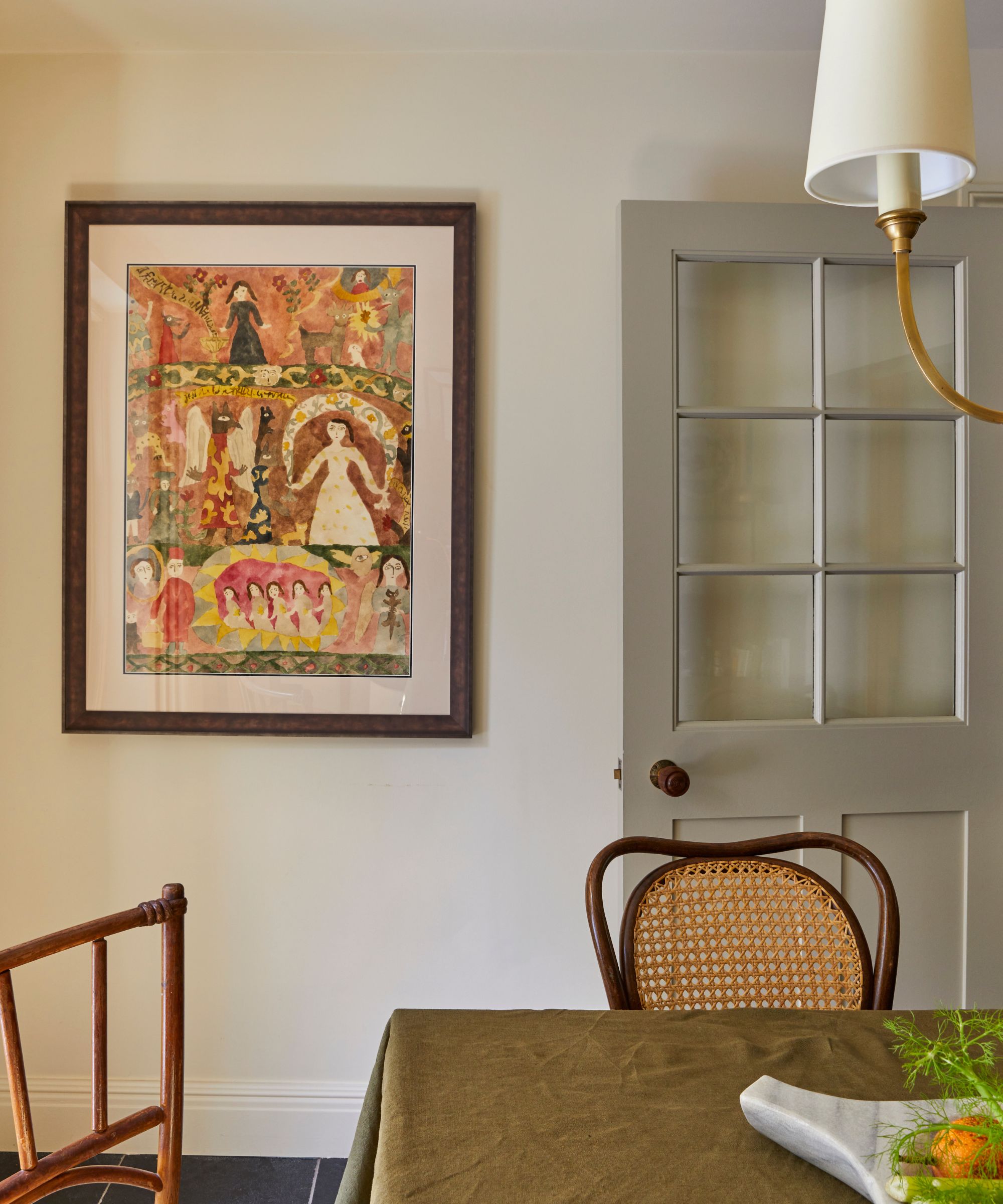 Dining room with a large folk print on the wall, a dining table with an olive green linen tablecloth, bistro dining chairs, an antler pendant light, and a grey painted glass door.