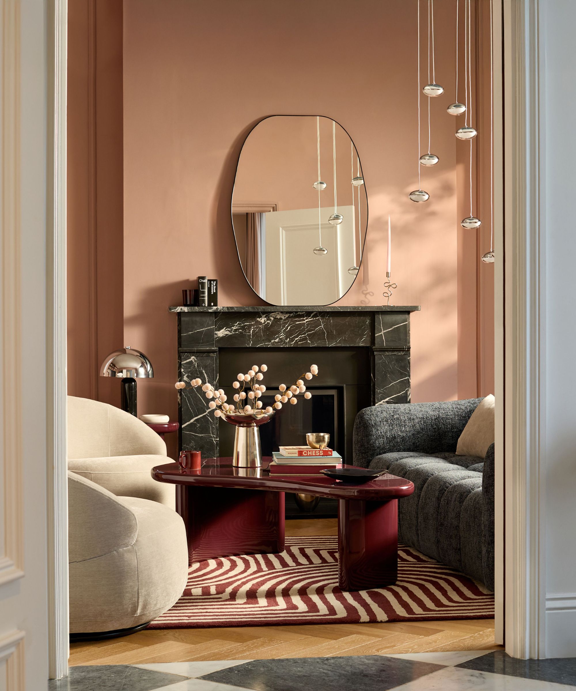 A terracotta pink living room with a dark marble fireplace, organic-shaped burgundy coffee table, cream curved sofa, grey boucl&amp;eacute; sofa and cascading glass pendant lights overhead.