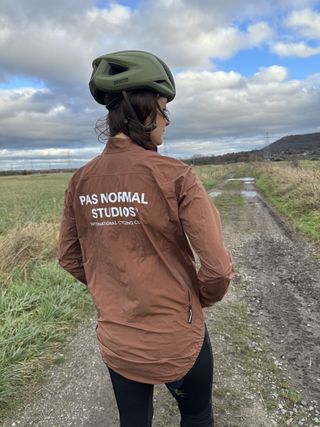 Woman in green helmet looks away from the camera, wearing a chocolate brown jacket with Pas Normal written on the back. She is stood in front of a muddy field.