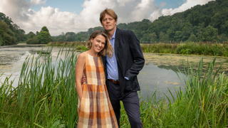Sally Bretton and Kris Marshall as Martha and Humphrey in Beyond Paradise, posing in front of a river and tall grass in a beautiful countryside setting