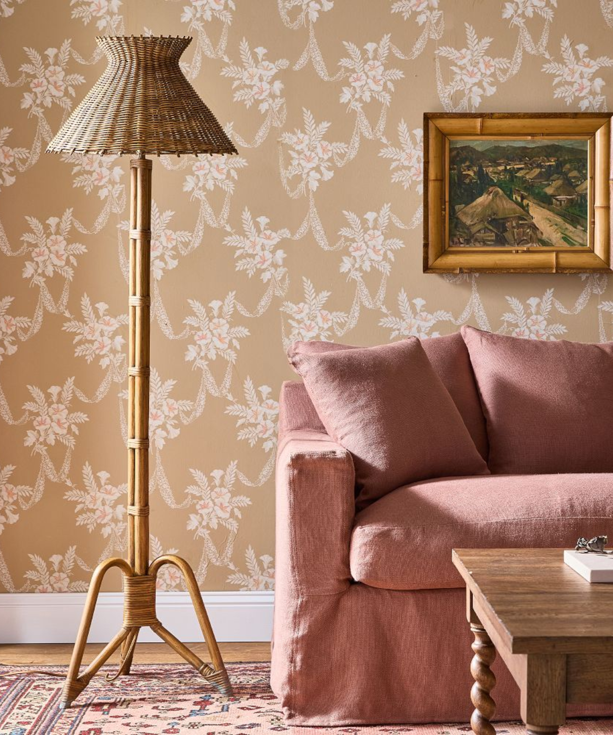 a room with tan floral wallpaper, a red slipcovered couch, a small artwork hanging from the wall, and a rattan floor lamp