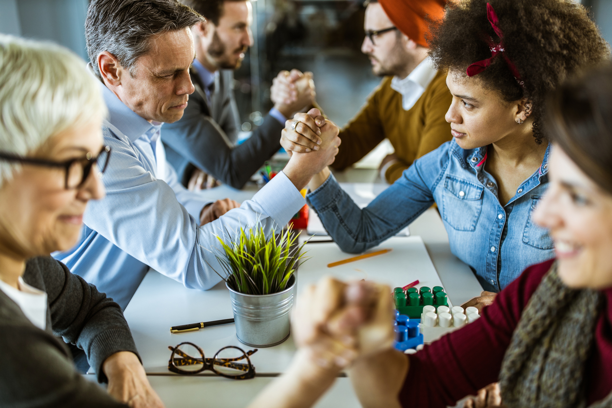A group of adults arm wrestles across a long table. The group is made up of all different kinds of people, including different ages.