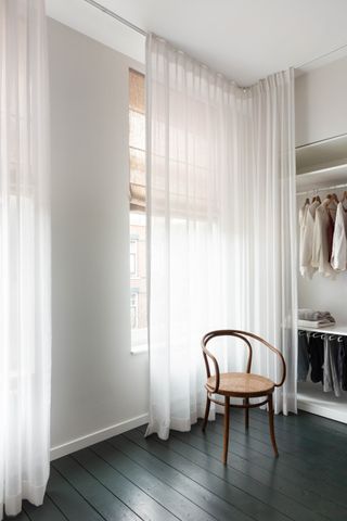 bedroom with black painted floorboards, rattan chair, and sheer curtains over windows that continue around a corner to cover the closet