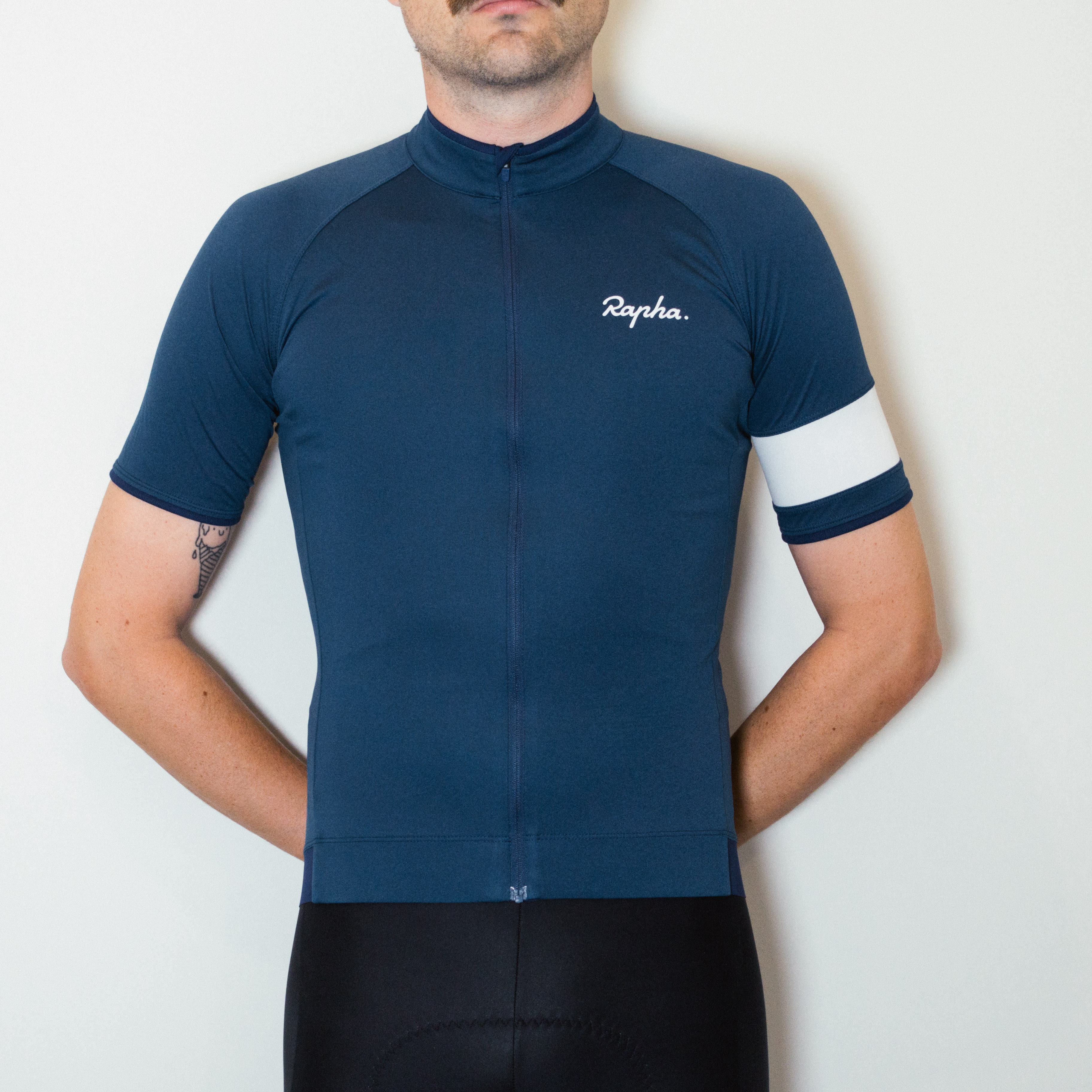 A white man in a blue cycling jersey and black cycling shorts stands against a white wall