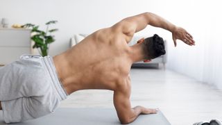Man with back to the camera and top off in a side plank with left arm extended overhead