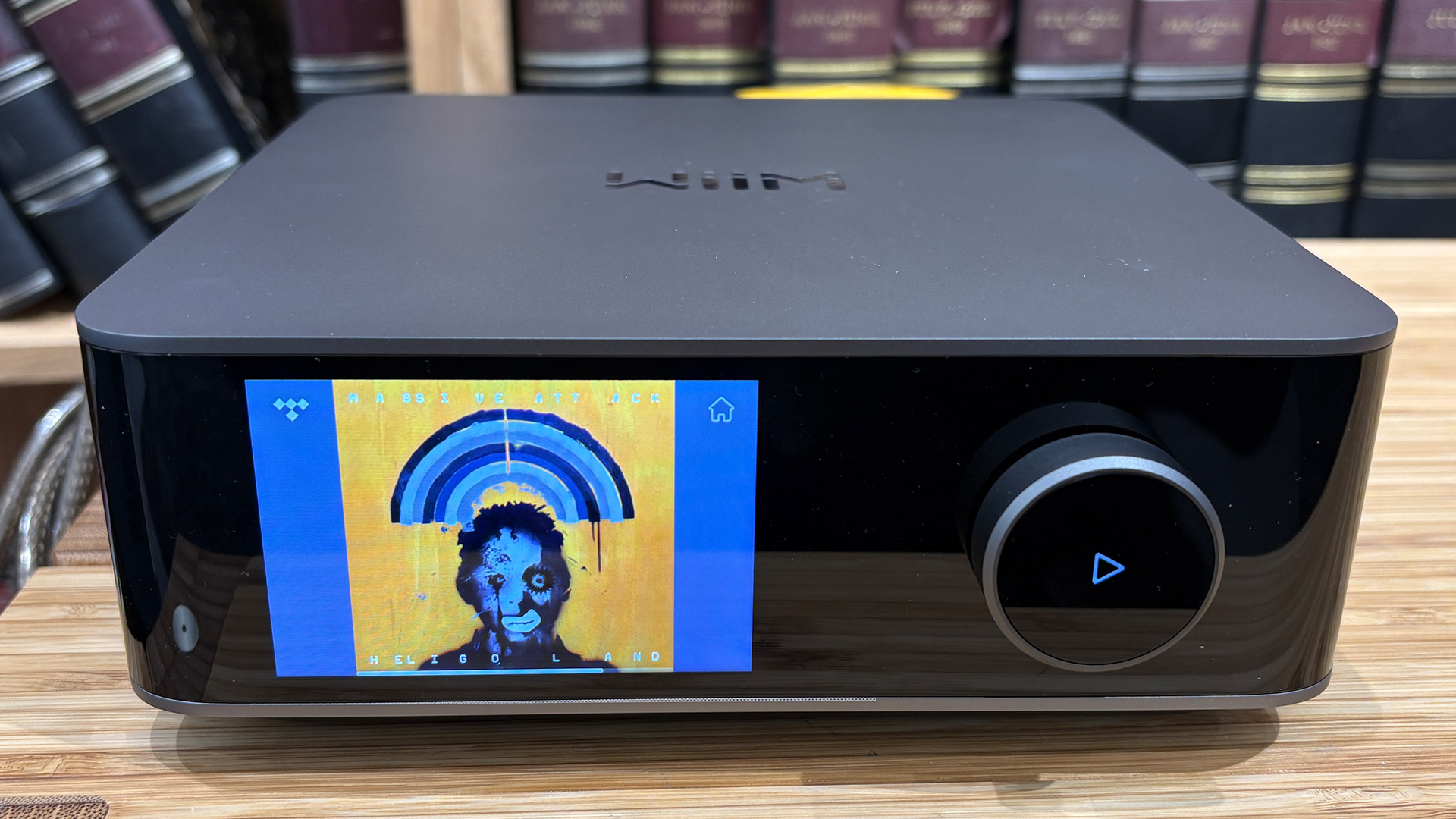 WiiM Amp Ultra streaming amplifier on wooden hi-fi rack in front of bookcase, on screen is album art