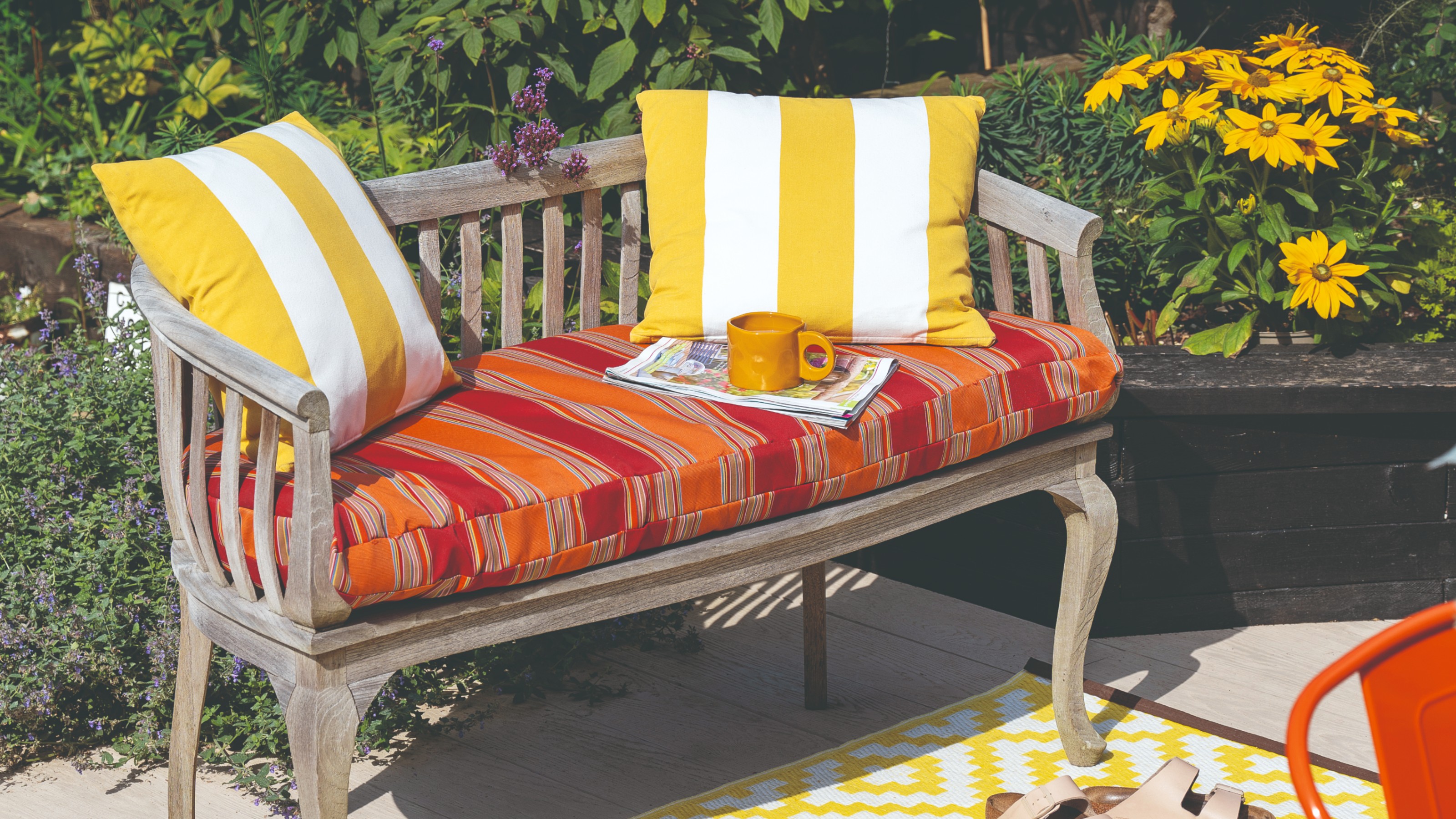 A sunny patio with a wooden garden bench with a striped seat cushion and deck chair-striped scatter cushions on top