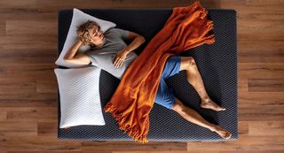 A man is seen from above, sleeping on the Layla Hybrid mattress with a bright orange blanket over his torso