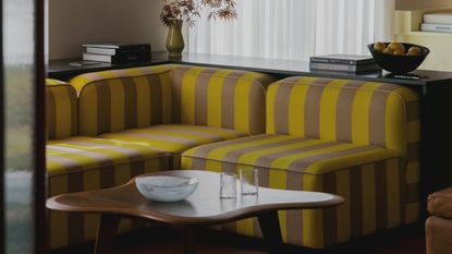 Image of a yellow and light brown/beige striped sofa with a low back in a living room. There is a console behind the sofa and an organically shaped coffee table in front of the sofa.
