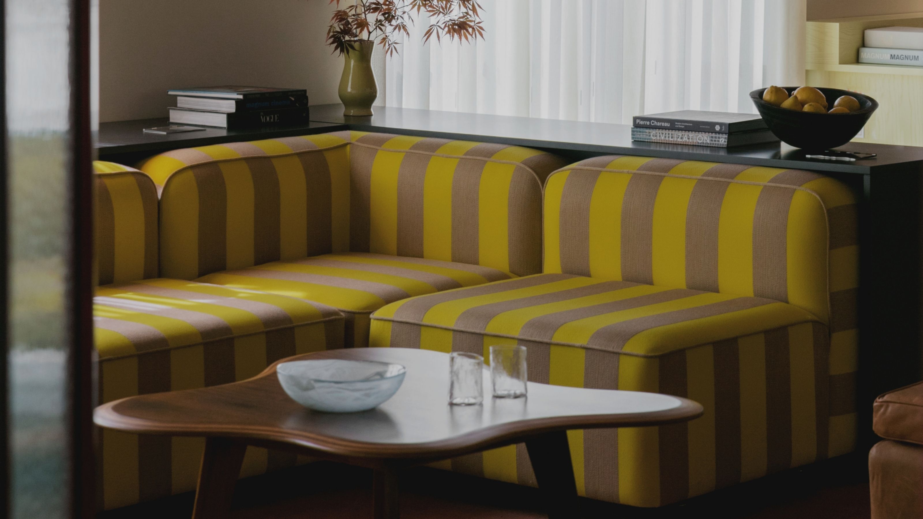Image of a yellow and light brown/beige striped sofa with a low back in a living room. There is a console behind the sofa and an organically shaped coffee table in front of the sofa.